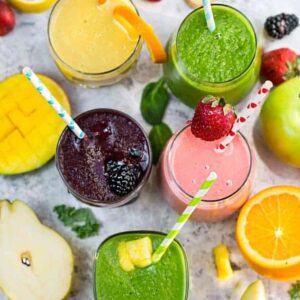 Top view of five healthy smoothies in a clear glass with a straw on a grey marble background with fruit