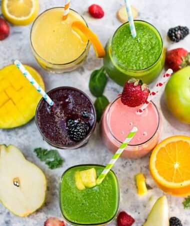 Top view of five healthy smoothies in a clear glass with a straw on a grey marble background with fruit