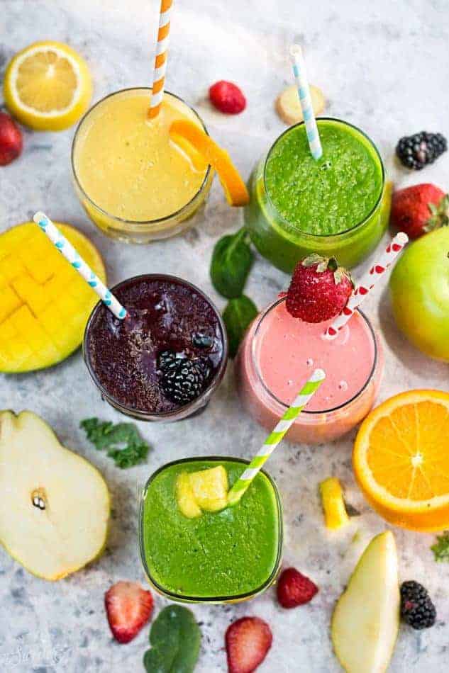 Top view of five healthy smoothies in a clear glass with a straw on a grey marble background with fruit