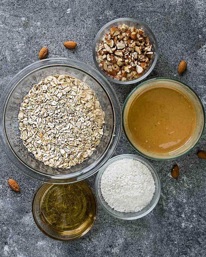 Overhead view of a bowl of oats, a bowl of chopped almonds, a bowl of shredded coconut, a bowl of almond butter, and a bowl of honey. 