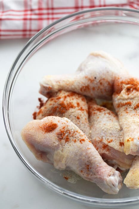 Six raw chicken drumsticks in clear mixing bowl with seasoning