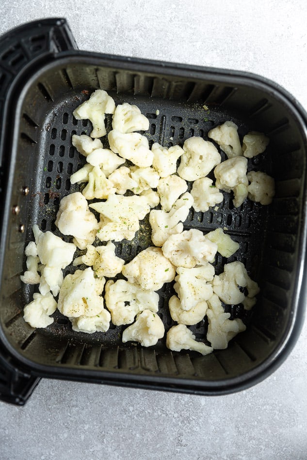Frozen cauliflower florets in black air fryer basket.