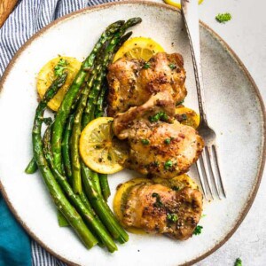 Three crispy air fryer chicken thighs on a white plate with a side of asparagus