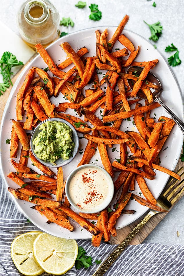 Top view of air fryer sweet potato fries on a white oval plate with avocado dip and cashew dip