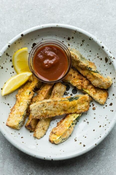 Top view of crispy air fryer zucchini Fries in a white bowl with lemon wedges and ketchup