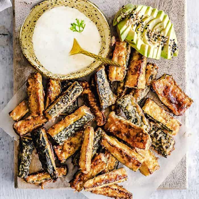 Top view of air fryer zucchini fries on a wooden cutting board and parchment paper with dip and avocado