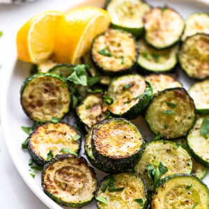A bunch of air-fried zucchini rounds on a white plate with two lemon wedges