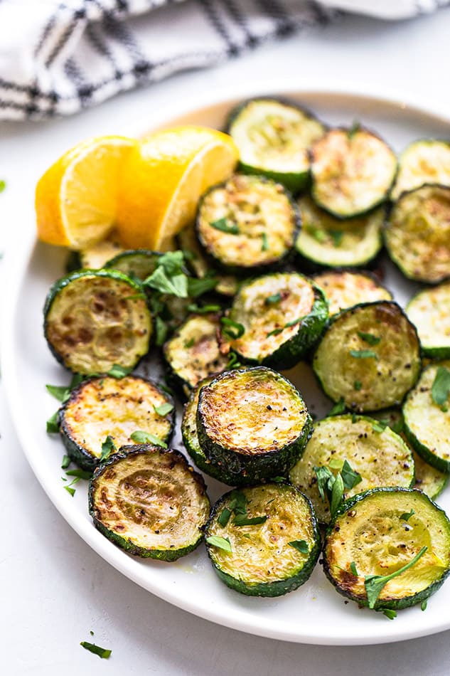 A bunch of air-fried zucchini rounds on a white plate with two lemon wedges