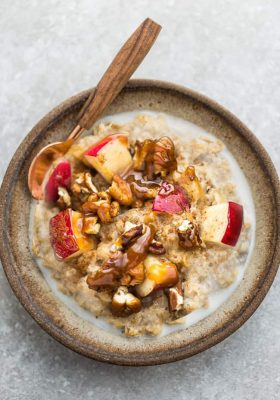 A bowl of apple oatmeal with a spoon topped with chopped apples, pecans, almond butter.