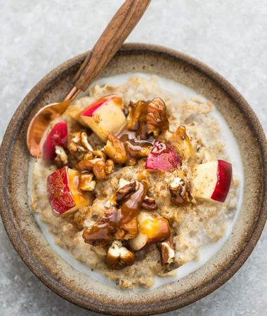 A bowl of apple oatmeal with a spoon topped with chopped apples, pecans, almond butter.
