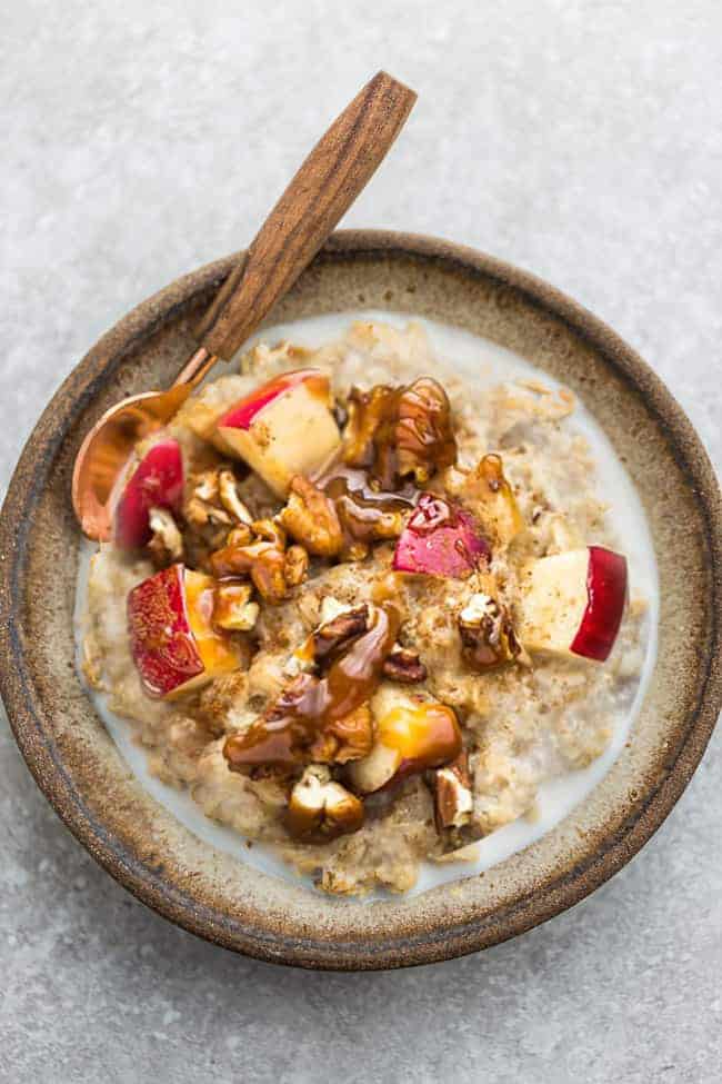 A bowl of apple oatmeal with a spoon topped with chopped apples, pecans, almond butter. 
