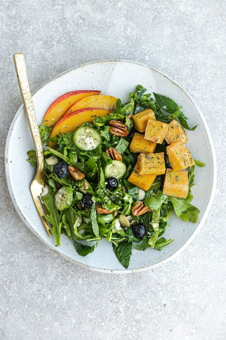 Top view of arugula salad with peaches, blueberries and air fryer tofu on a white plate on a grey background