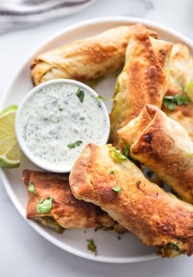 Overhead shot of 8 crispy avocado egg rolls on a white plate with a serving of cilantro dipping sauce