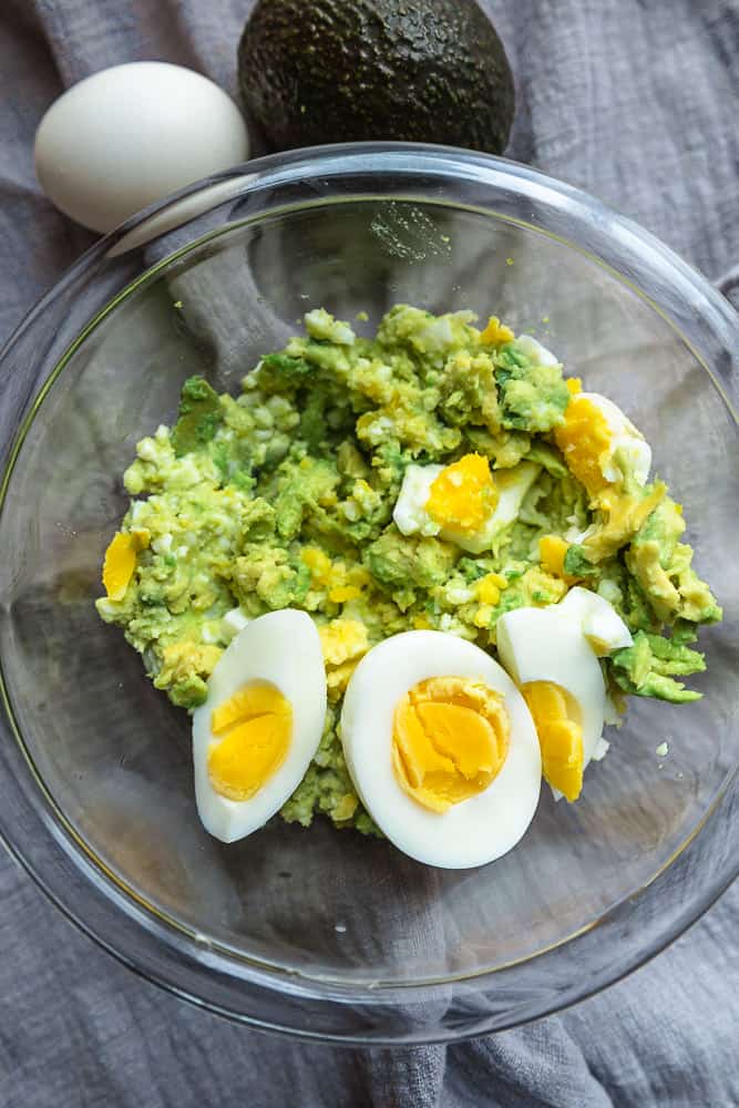 A keto avocado egg salad in a clear mixing bowl with extra hard boiled egg halves on top
