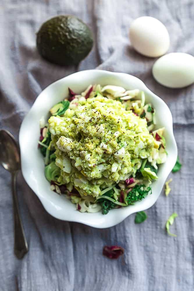 A bowl filled with keto egg salad beside a spoon and two eggs