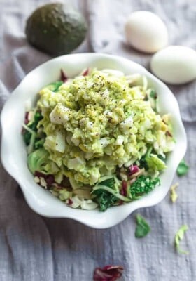 A bowl of avocado egg salad with two hard-boiled eggs and an avocado in the background