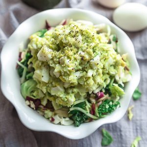 A bowl of avocado egg salad with two hard-boiled eggs and an avocado in the background