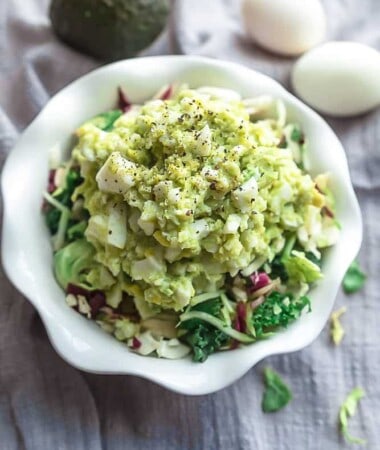 A bowl of avocado egg salad with two hard-boiled eggs and an avocado in the background