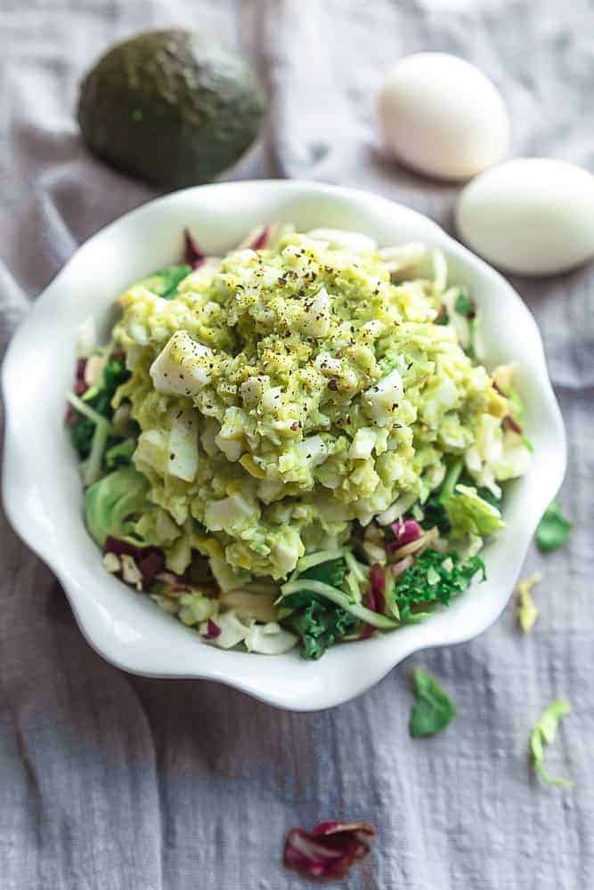 A bowl of avocado egg salad with two hard-boiled eggs and an avocado in the background