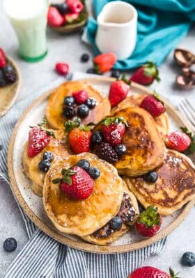 Top view of six keto coconut flour pancakes on a plate with strawberries, blueberries and sugar free syrup on a grey background