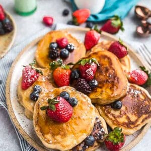 Top view of six keto coconut flour pancakes on a plate with strawberries, blueberries and sugar free syrup on a grey background