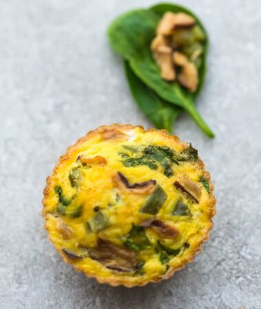 A Bacon, Mushroom and Spinach Breakfast Egg Muffin Beside Two Spinach Leaves and a Piece of Mushroom