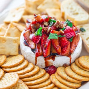 Side shot of a pile of Balsamic Strawberries piled on baked brie served with an assortment of gluten-free crackers and toasted baguettes