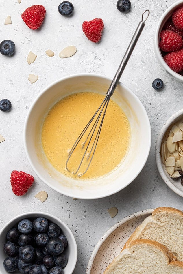 A white bowl containing french toast custard with a whisk inside on a white background, surrounded by berries.