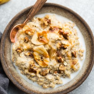 A serving of banana oatmeal in a bowl with walnuts and a banana behind it