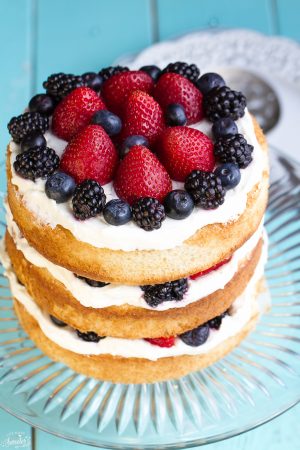 Simple Berries & Cream Sponge Cake topped with blueberries, blackberries and strawberries on a glass cake platter.