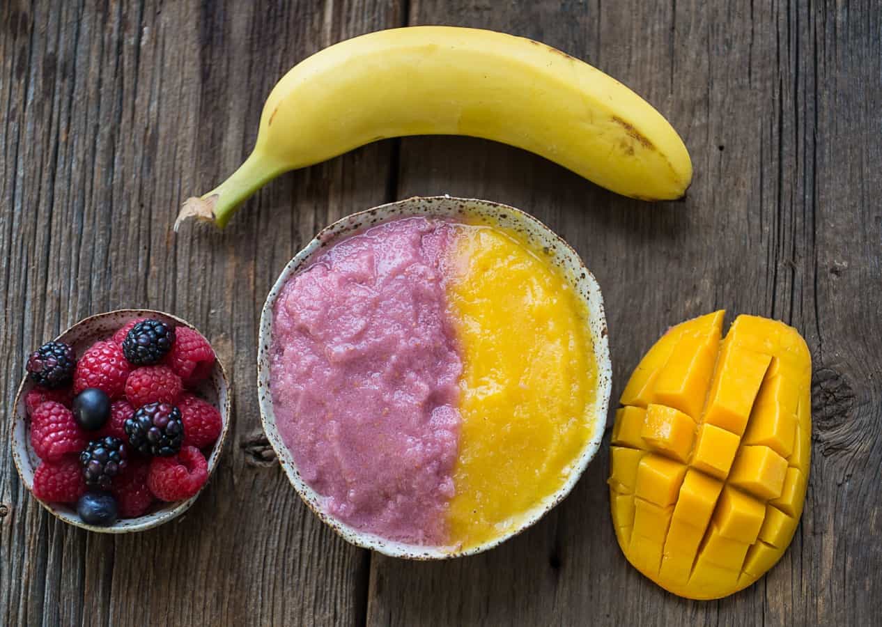 Close-up image of orange and pink berry mango smoothie bowl paired with fresh mango, banana, and berries.