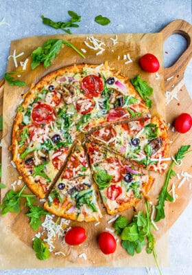 Top view of cauliflower pizza crust on parchment paper with arugula, pepperoni and tomatoes