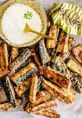 Top view of crispy baked zucchini fries on parchment paper with a bowl of dip and avocado slices