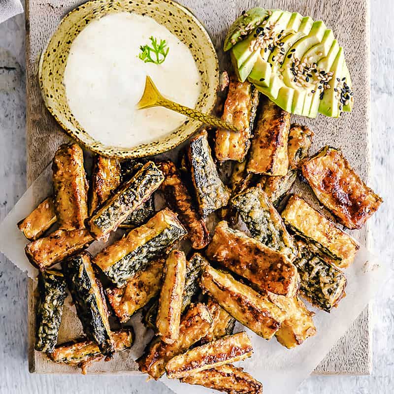 Top view of crispy baked zucchini fries on parchment paper with a bowl of dip and avocado slices