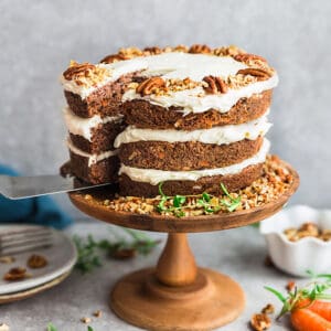 Side view of gluten free Keto Carrot Cake on wooden cake stand with chopped pecans and carrots