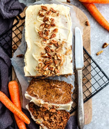 Top view of a paleo carrot cake loaf with two slices on a cutting board on a grey background with a knife and carrots on the side