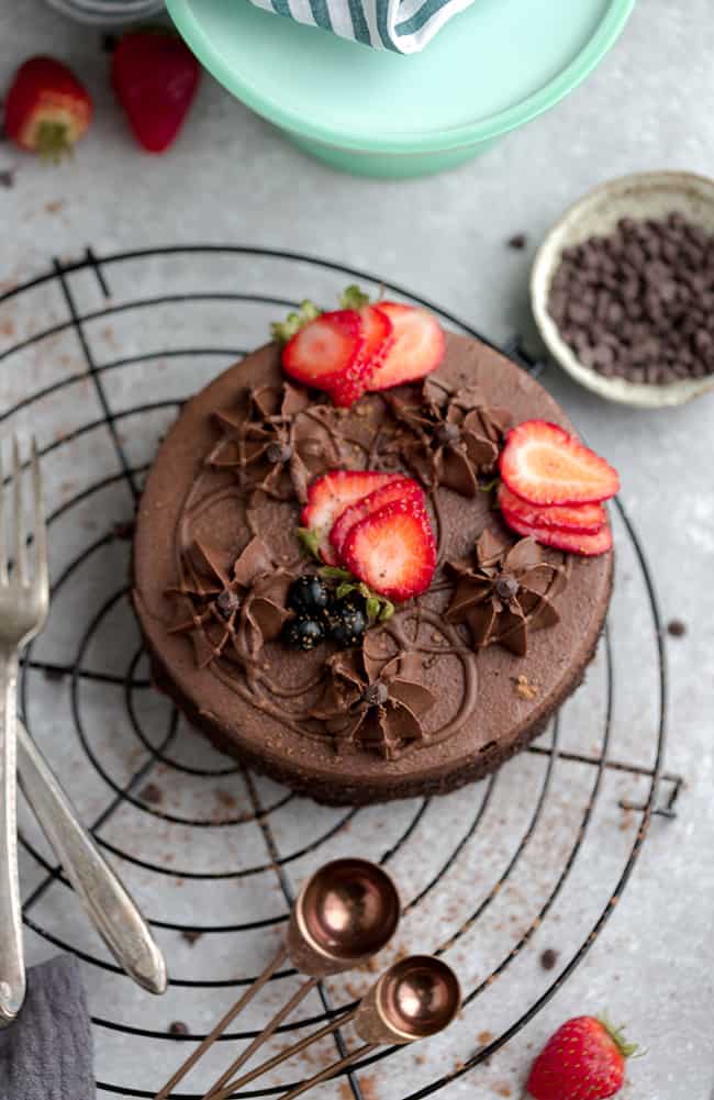 Top view of a whole keto chocolate cake on a wire rack and a grey background with strawberries and sugar free frosting