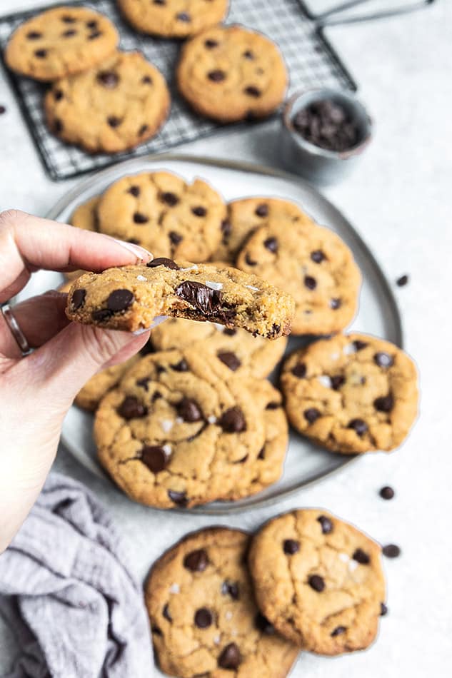 A hand holding a Vegan Chocolate Chip Cookie with a bite out of it and a plate of cookies in the background