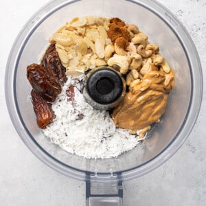 Top view of dates, cashews, coconut, nut butter, cinnamon, protein powder and vanilla in a food processor