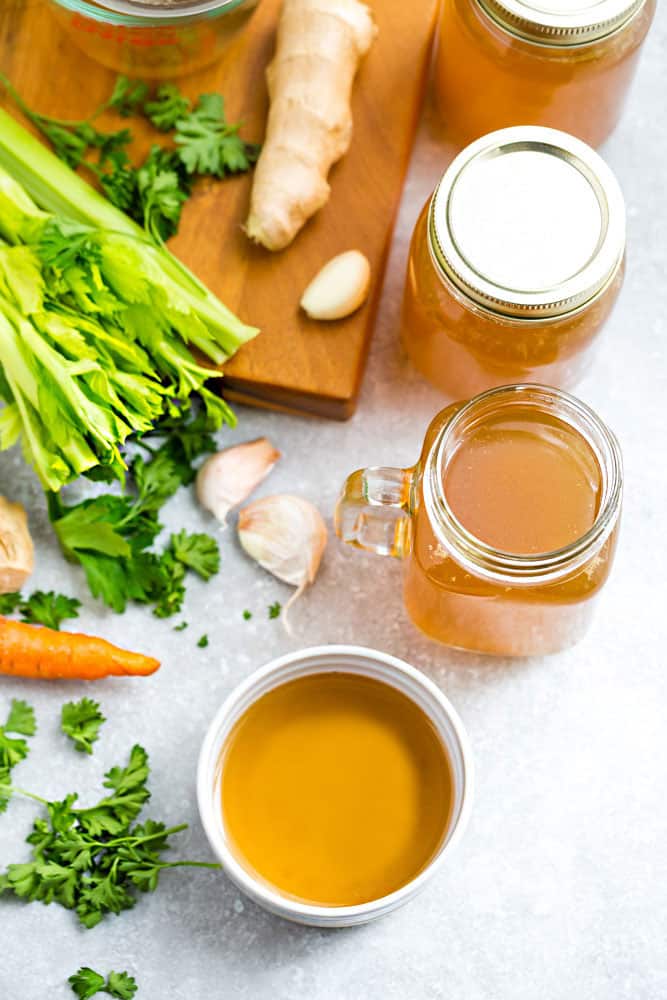 Glass jars of homemade Bone Broth surrounded by ginger, celery, garlic and carrots.