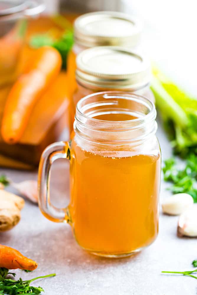 Three glass jars of Instant Pot Bone Broth surrounded by fresh vegetables.