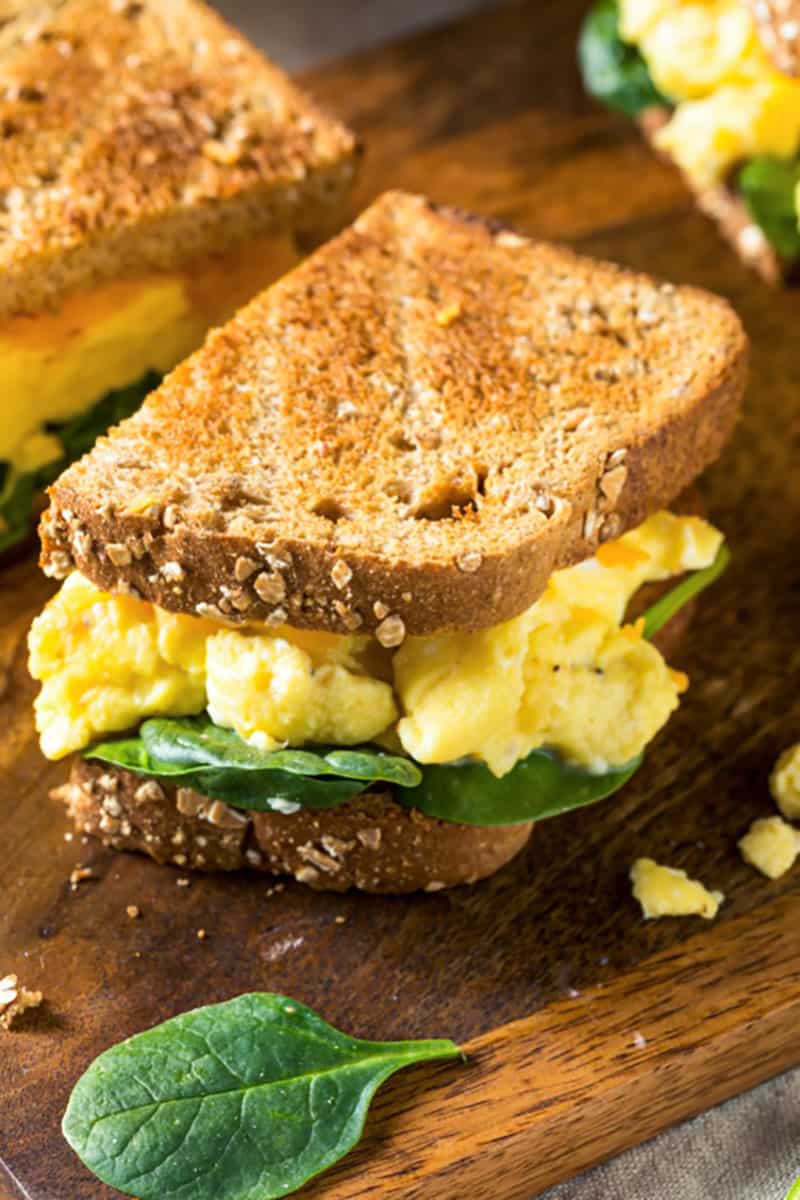 A breakfast sandwich cut in half, with pieces of spinach and scrambled eggs falling out 