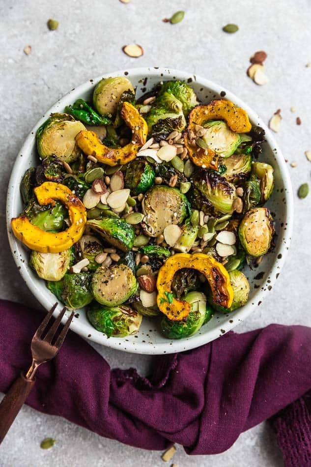 Top view of cooked Brussels sprouts and delicata squash and pumpkin seeds in a white bowl and a copper fork