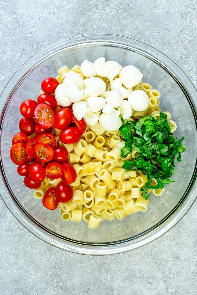 Top view of pasta, tomatoes, mozzarella, and fresh basil in glass bowl on grey surface for caprese pasta salad.
