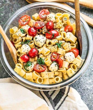 Top view of gluten free caprese pasta salad in a clear mixing bowl with two wooden spoons