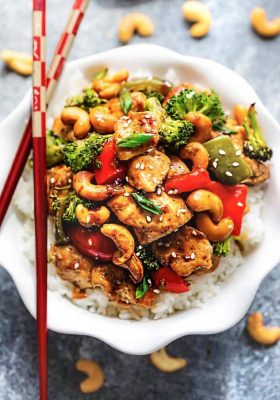 Cashew chicken on a bed of white rice inside of a bowl with a pair of chopsticks balancing on the rim