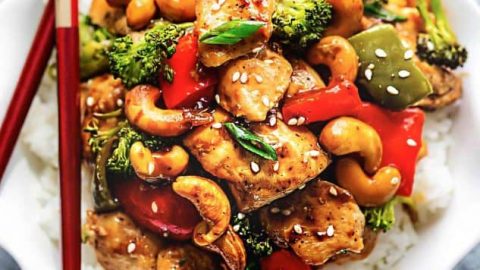 Cashew chicken on a bed of white rice inside of a bowl with a pair of chopsticks balancing on the rim