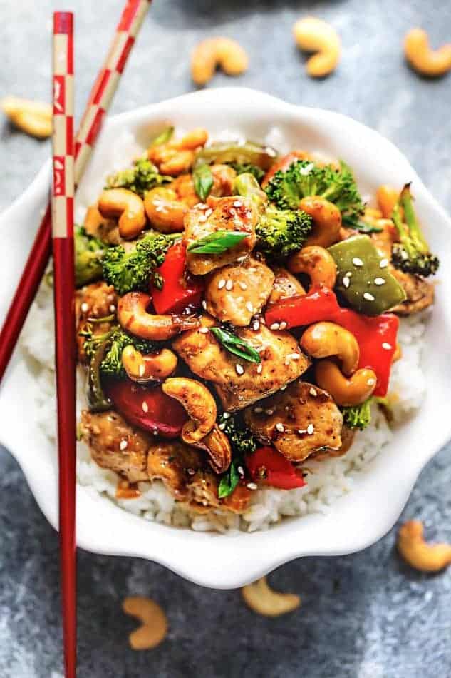 Gluten free Cashew Chicken on a bed of white rice in a small white bowl with red chopsticks.