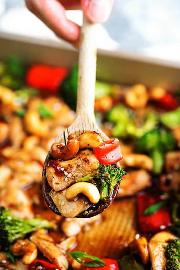 A scoop of Cashew Chicken on a wooden spoon over a sheet pan