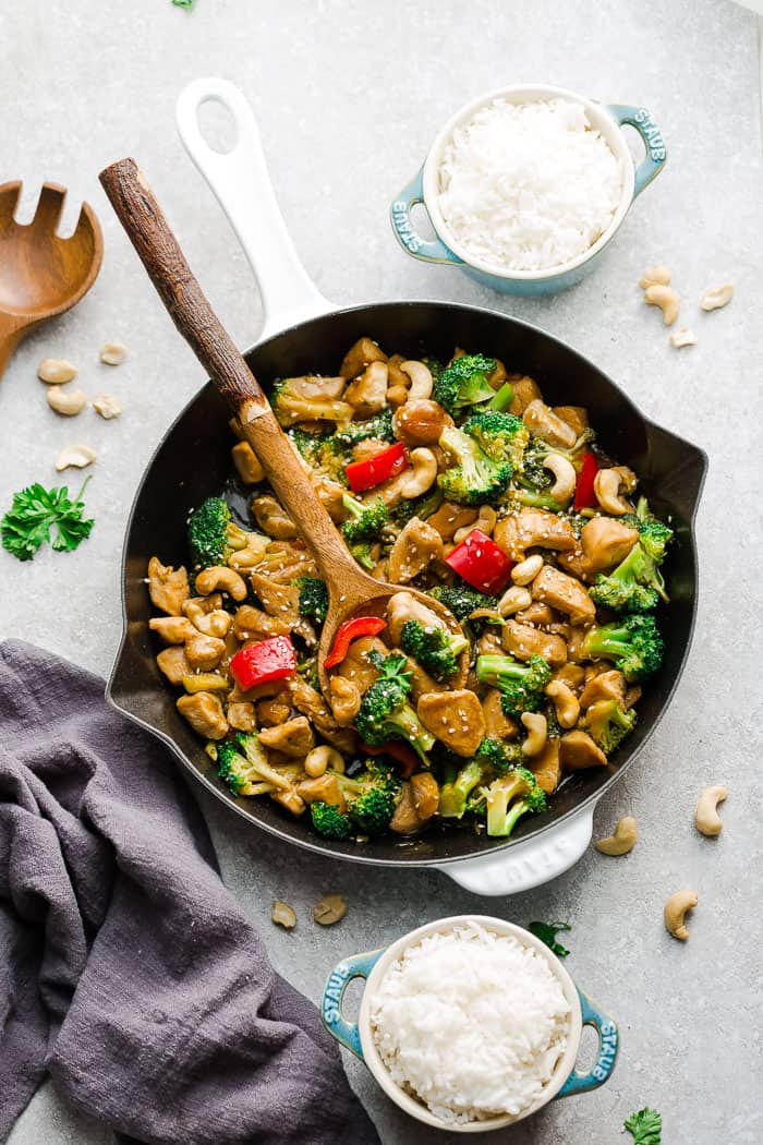 A cashew chicken stir fry inside of a skillet with two bowls of rice beside it